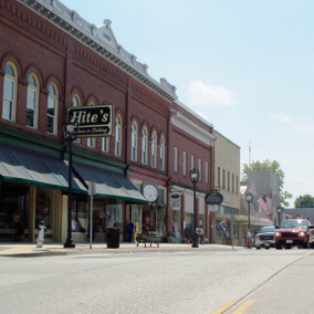A street with shops in the historic area of downtown Clarksville, VA. There are cars driving down the road, and a sign hanging from a building that reads, “Hite’s Fashion in Clothing.”