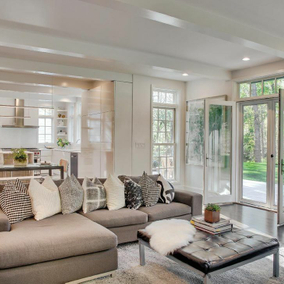 Home featuring a universal design. Living room with a gray sofa, neutral-colored pillows, a leather ottoman and a gray rug. There is a kitchen behind the living room.